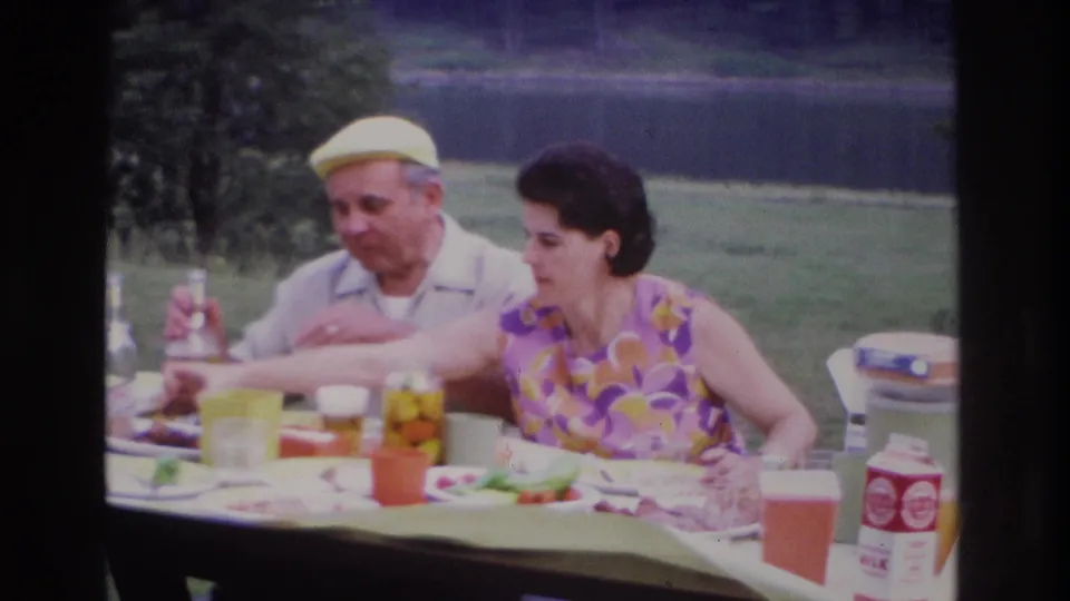 1967 Ottawa lakeside picnic frame showing a woman reaching across the table as an elderly man watches, with food and cartons visible.