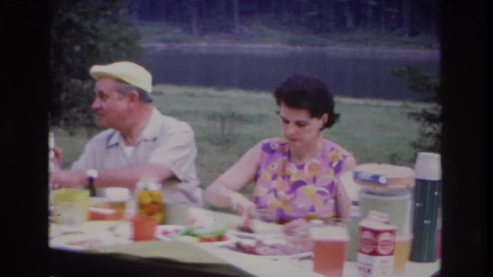 1967 Ottawa home-movie frame of an elderly man gesturing during a picnic as the woman looks down at the table.