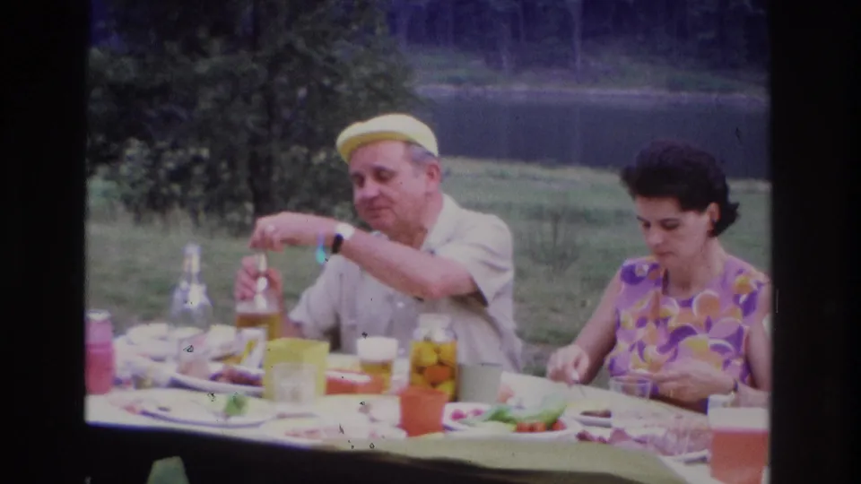 1967 Ottawa lakeside picnic frame showing the man reaching toward the table while the woman eats beside jars and plates.