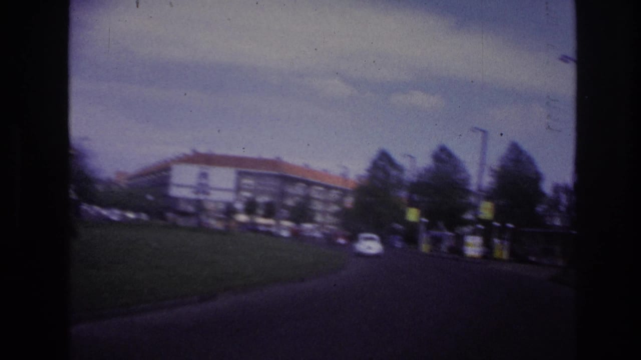 1967 Paris driving view past a grassy verge, apartment blocks, trees, and a small white car on a curved urban road.