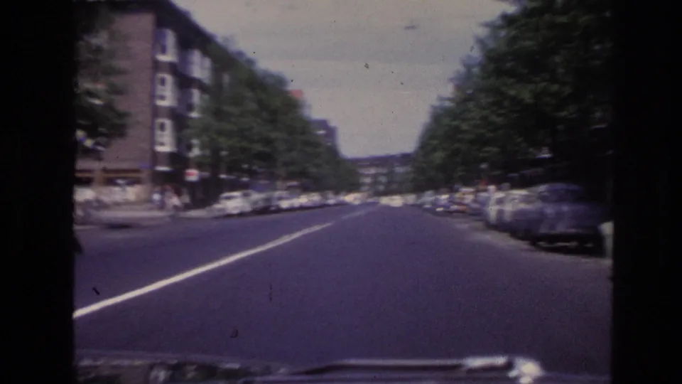1967 Paris driving POV down a tree-lined street with parked vintage cars, apartment facades, and small pedestrians.