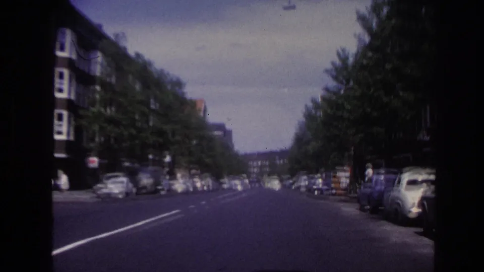 1967 Paris street view from a moving vehicle, showing parked cars, trees, apartment buildings, and distant road activity.