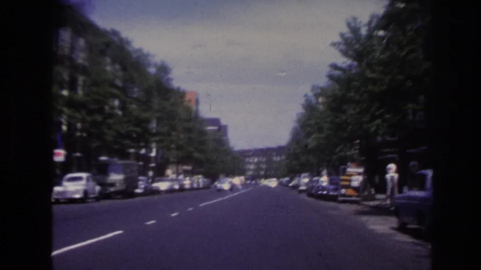 1967 Paris street view from a moving vehicle, showing a broad road, parked cars, trees, and apartment buildings.