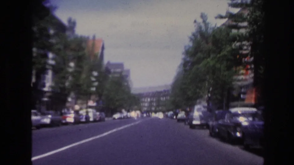 Soft 8mm frame of a 1967 Paris street, with parked cars, a broad roadway, trees, and distant buildings.