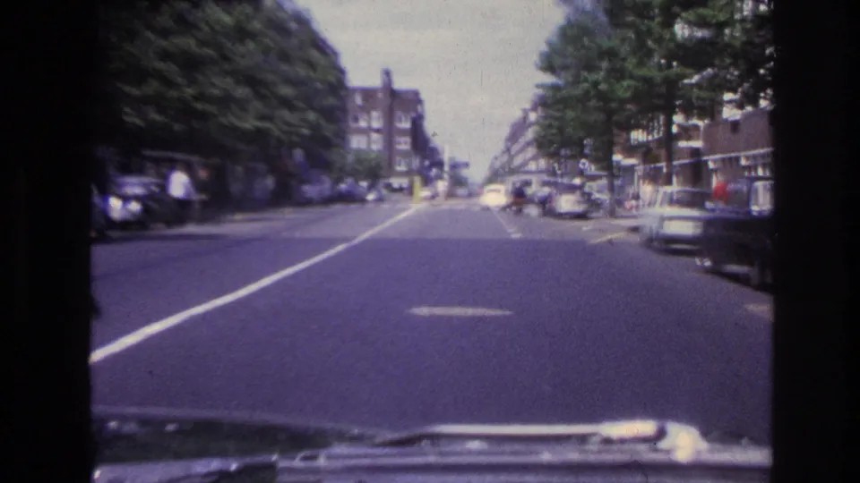 1967 Paris driving shot with the car hood visible at the bottom, parked cars along the street, trees, and apartment blocks.
