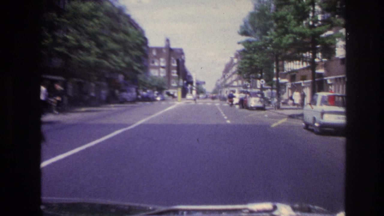 1967 Paris home-movie frame from a vehicle approaching a tree-lined street with parked cars and apartment blocks.