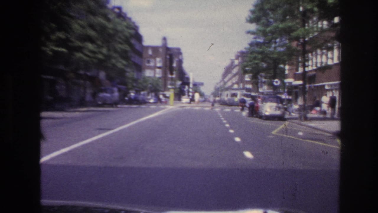 Forward road view with parked cars, trees, buildings, and pedestrians near the curb.