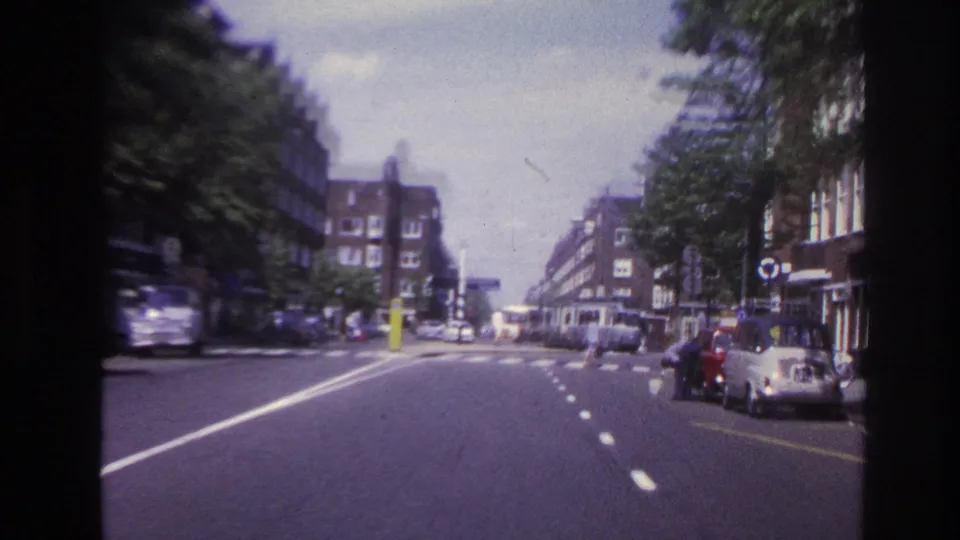 1967 Paris moving-vehicle frame showing a tree-lined roadway, curbside cars, pedestrians, and mid-century apartment blocks.