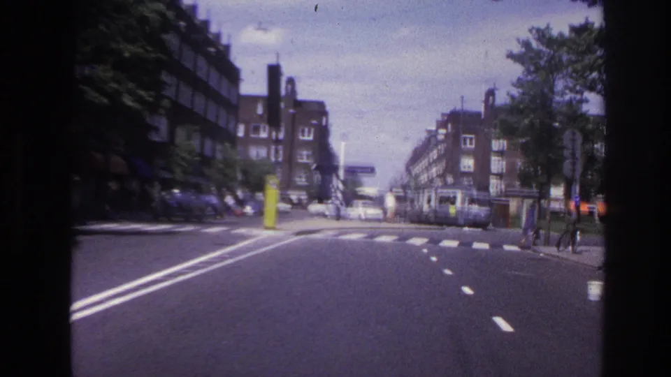 1967 Paris home-movie frame approaching a crosswalk or junction with pedestrians, parked cars, trees, and apartment buildings.