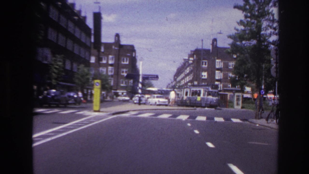 1967 Paris street corner from a moving vehicle, with roadway markings, mid-century cars, a tram or streetcar, and apartment buildings.