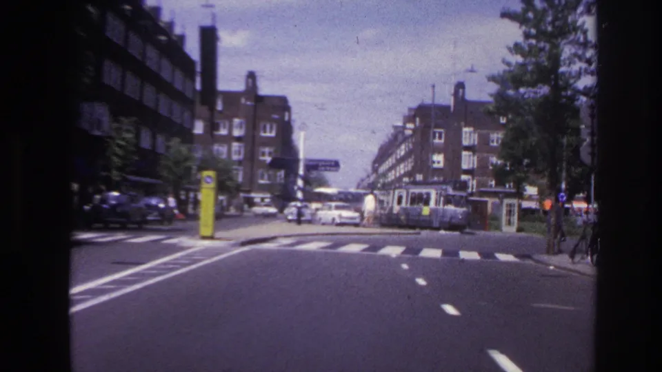 1967 Paris street corner from a moving vehicle, with roadway markings, mid-century cars, a tram or streetcar, and apartment buildings.