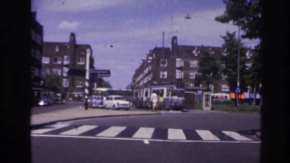 Moving vehicle view in 1967 Paris showing a crosswalk, street traffic, a tram or streetcar, and multi-story buildings beyond the corner.