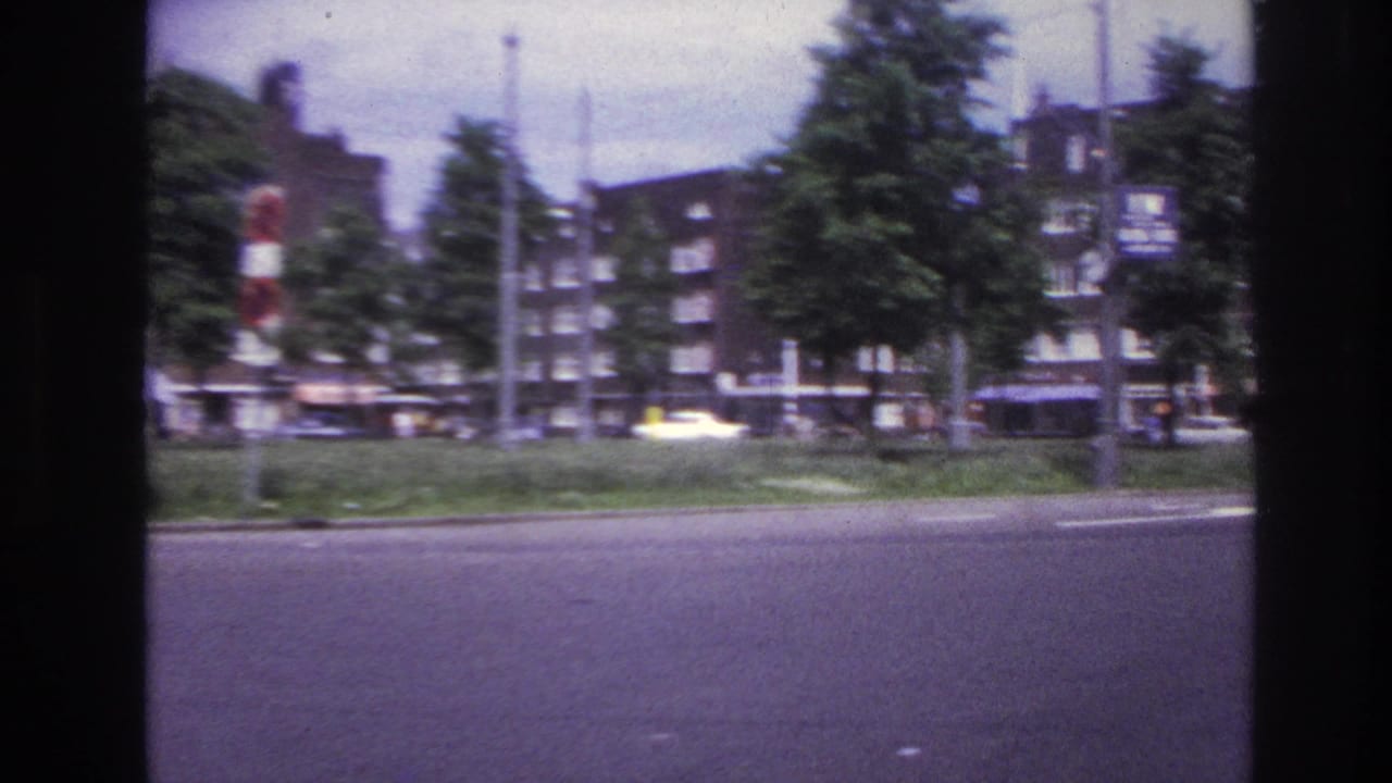 1967 Paris home-movie frame showing a broad roadway bordered by trees and multi-story apartment buildings, with small cars and traffic signals visible.