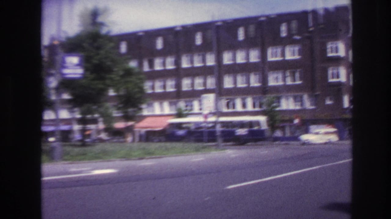 Urban road with apartment block facades, storefront awnings, roadside trees, and cars near an intersection.