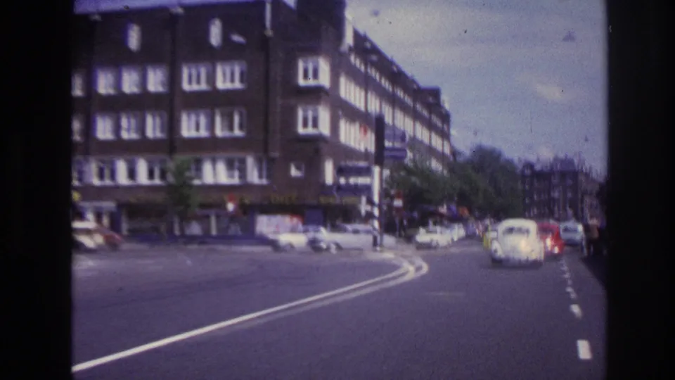 1967 Paris road scene from a moving vehicle, with a pale compact car ahead on a curve and apartment blocks framing the street.