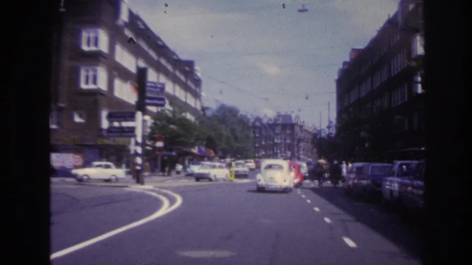 1967 Paris traffic frame showing a vehicle-eye view along a curved road, with compact cars, parked vehicles, trees, and apartment buildings.