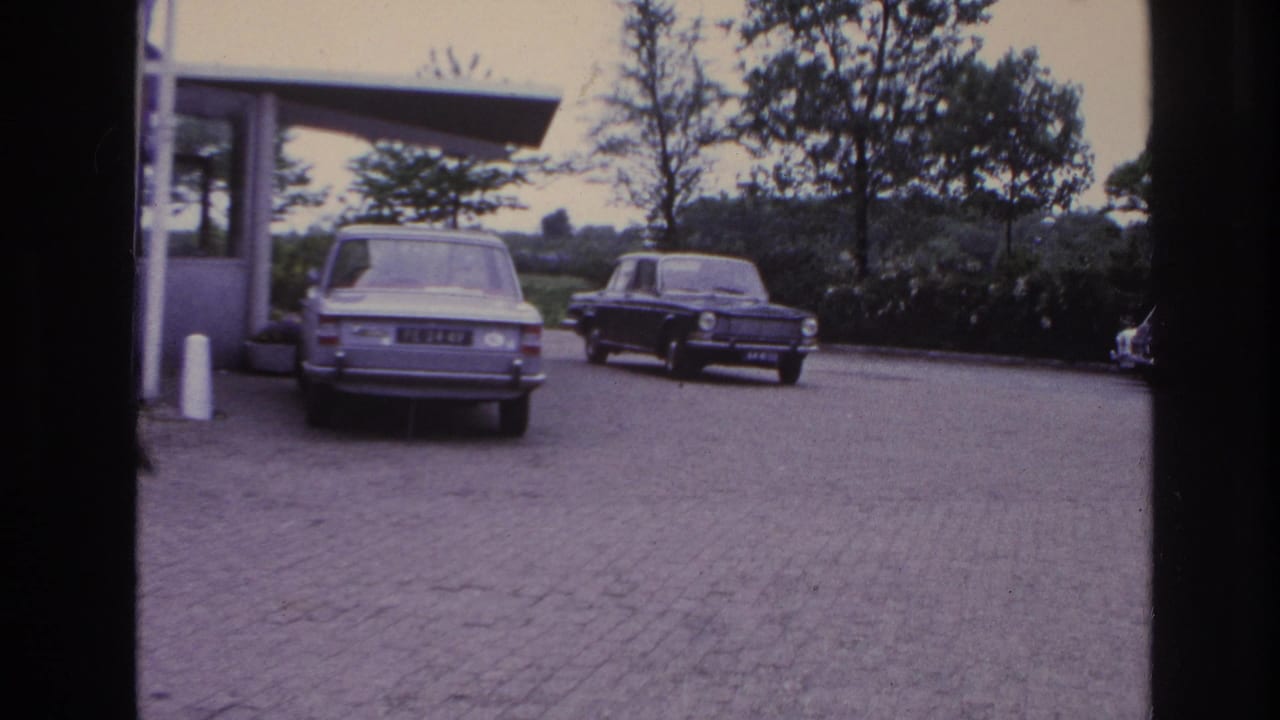 1967 Paris parking-area view with a light sedan, dark car, cobblestone surface, flat-roof structure, and trees in the background.