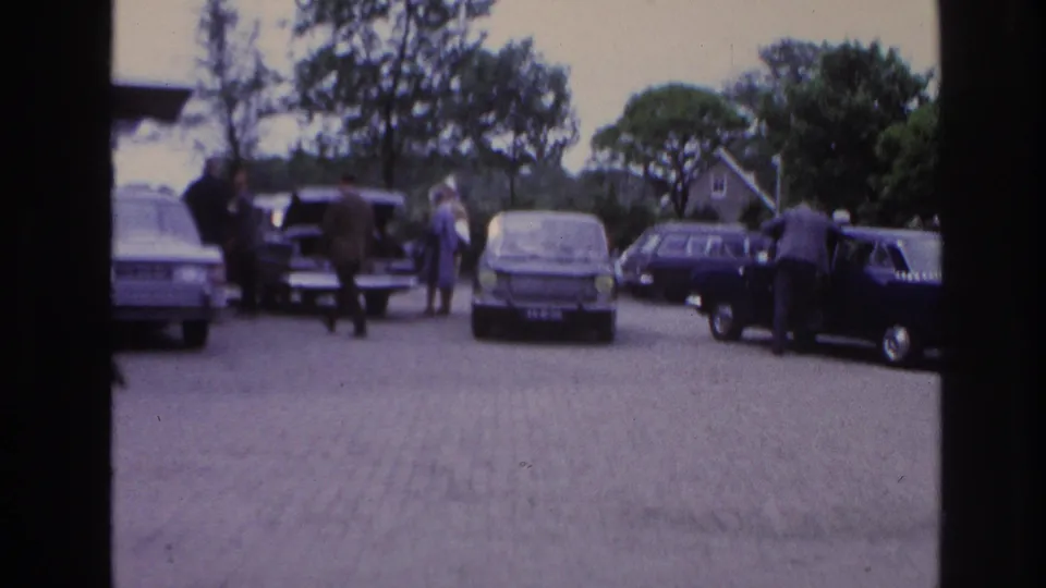 1967 Paris parking-area view with several classic cars, adults near the vehicles, cobblestone paving, trees, and soft handheld 8mm focus.