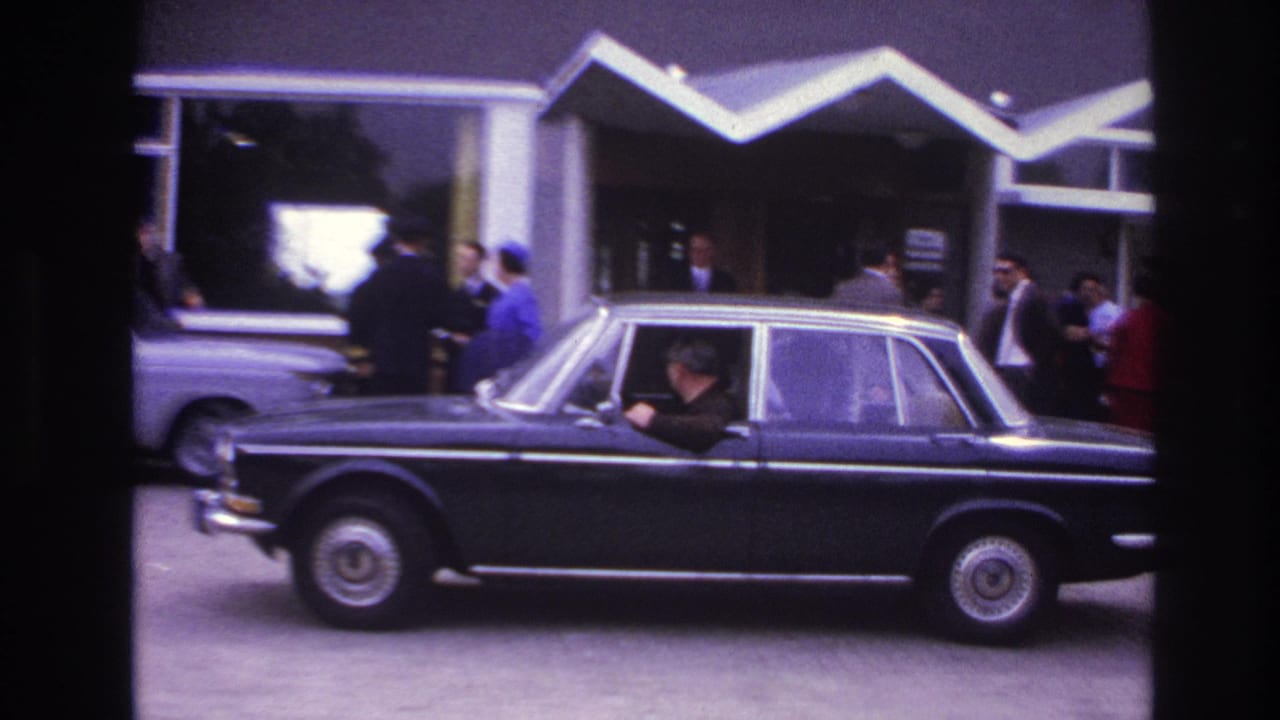 Black sedan passing a modernist entrance with people gathered near the doors.