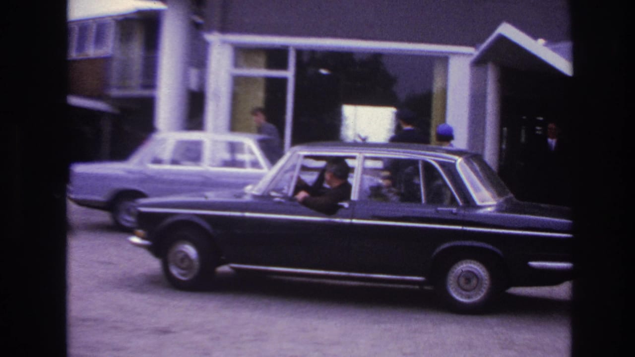 Black sedan with driver and rear passengers moving across a Paris building forecourt in 1967 footage.