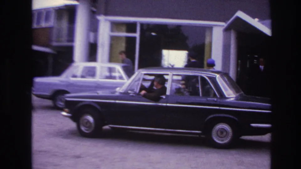 Black sedan with driver and rear passengers moving across a Paris building forecourt in 1967 footage.