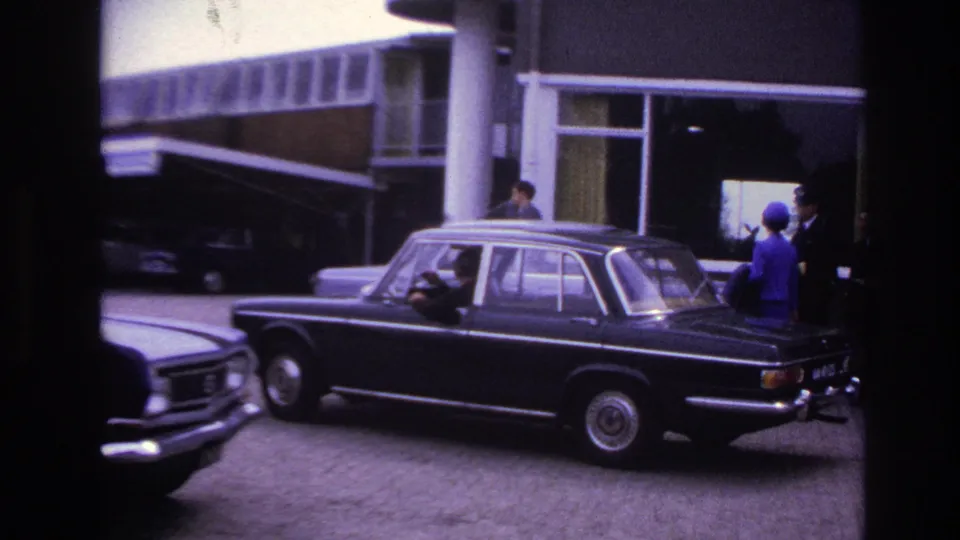 Rear three-quarter view of the black sedan turning or passing near a modernist entrance in 1967 Paris footage.
