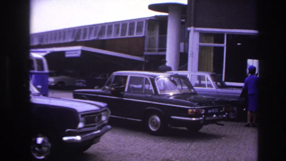 Black sedan moving away from a Paris modernist entrance, with period cars and a person in blue visible nearby.