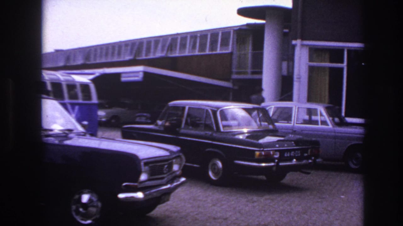 1967 Paris home-movie frame showing classic cars in a motel parking area, with a blue-and-white camper van and modern facade behind them.
