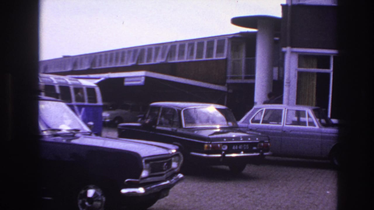 Classic cars remain parked outside the motel in this 1967 Paris travel frame, with the building frontage and camper van visible in the background.