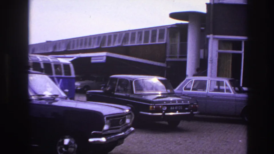 Classic cars remain parked outside the motel in this 1967 Paris travel frame, with the building frontage and camper van visible in the background.