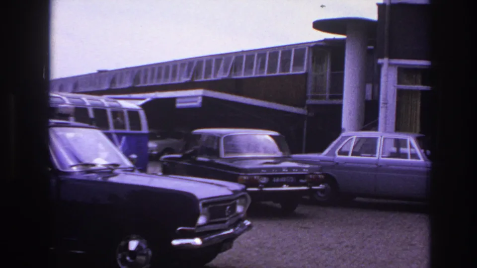 A handheld 8mm view shows classic sedans and a camper van outside the Paris motel, with the building's windows and entrance area behind them.