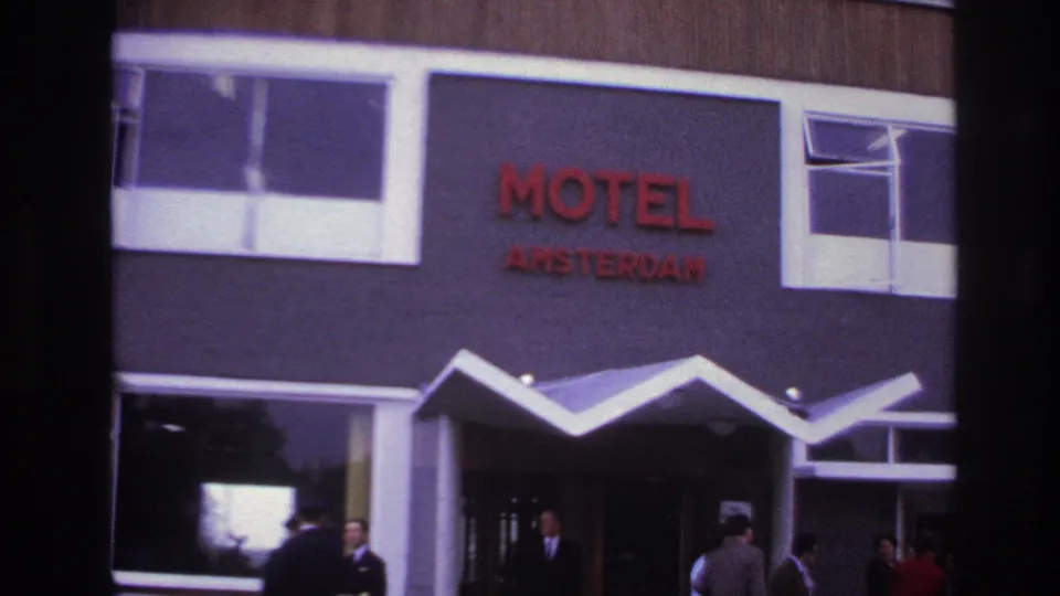 1967 Paris home-movie frame showing the Motel Amsterdam facade, red lettering, white zigzag awning, people near the entrance, and a light-colored car.