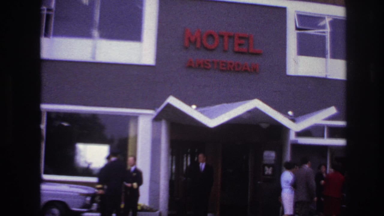 Closer view of Motel Amsterdam entrance with staff, guests, red sign, awning, and parked car.