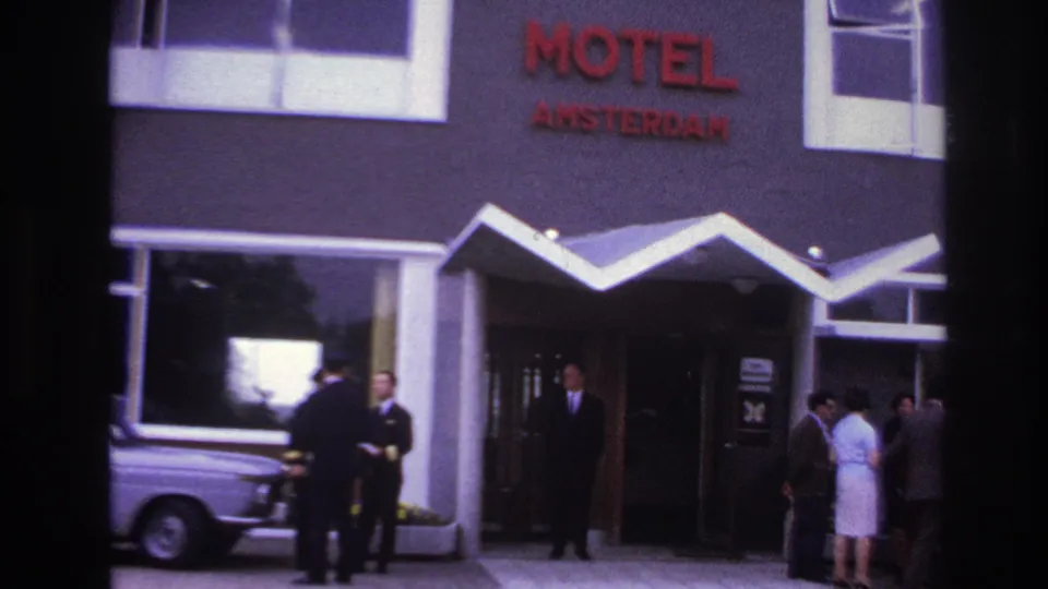 1967 Paris home-movie still showing guests and staff outside Motel Amsterdam, with a pale-dressed woman, suited men, signage, awning, and a car.