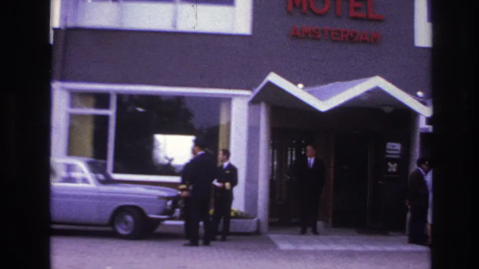 1967 Paris frame of the Motel Amsterdam exterior, showing the red sign, white awning, staff at the doorway, guests at right, and a light-colored car.