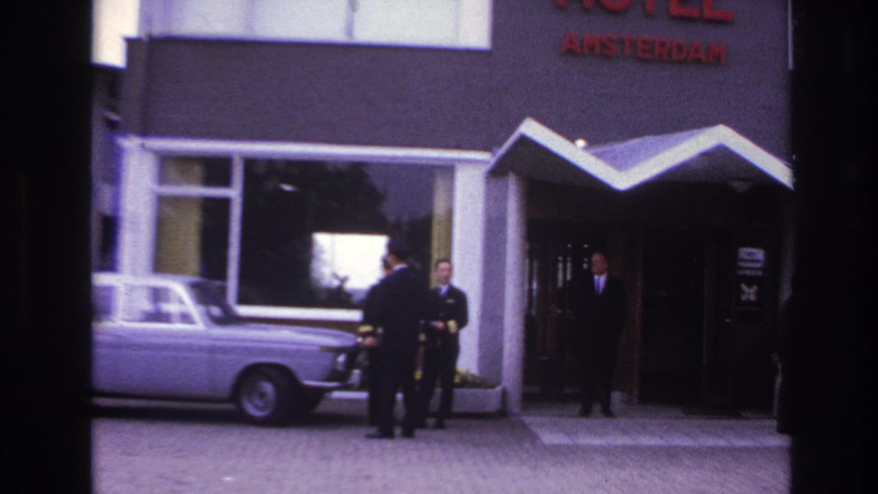 1967 Paris hotel exterior with suited men waiting near a canopy entrance and a pale sedan under a red HOTEL AMSTERDAM sign.