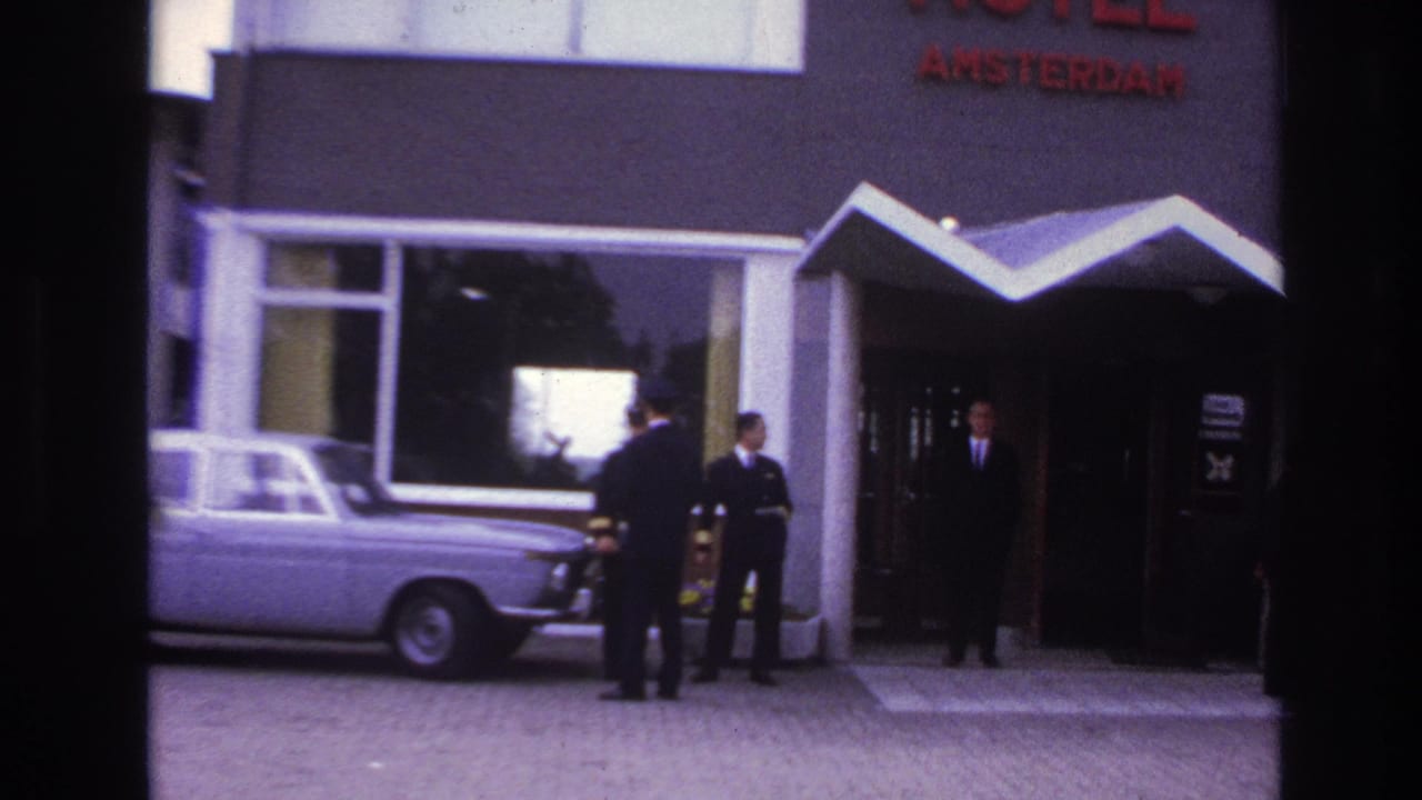 Suited men wait and face one another outside the Paris hotel entrance, with the red sign and pale sedan visible.