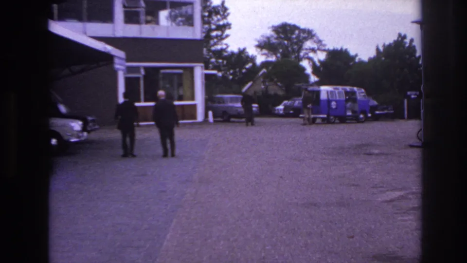 1967 Paris hotel forecourt with two suited men walking toward cars and a blue and white van in the background.