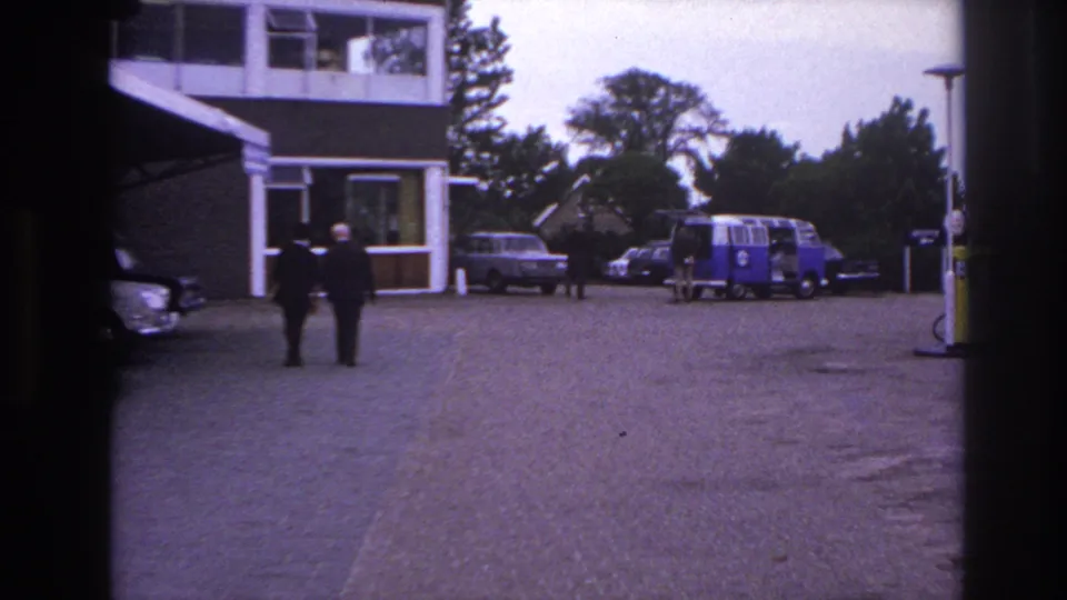 Wide 1967 Paris hotel exterior showing suited men in the forecourt, parked cars, trees, and a blue and white van.