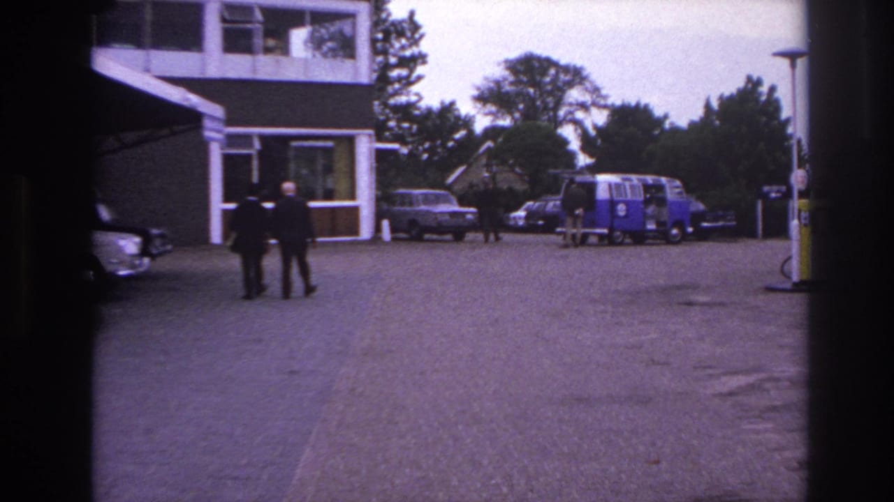 1967 Paris home-movie frame of two suited men walking toward a modernist entrance with classic cars and a van nearby.