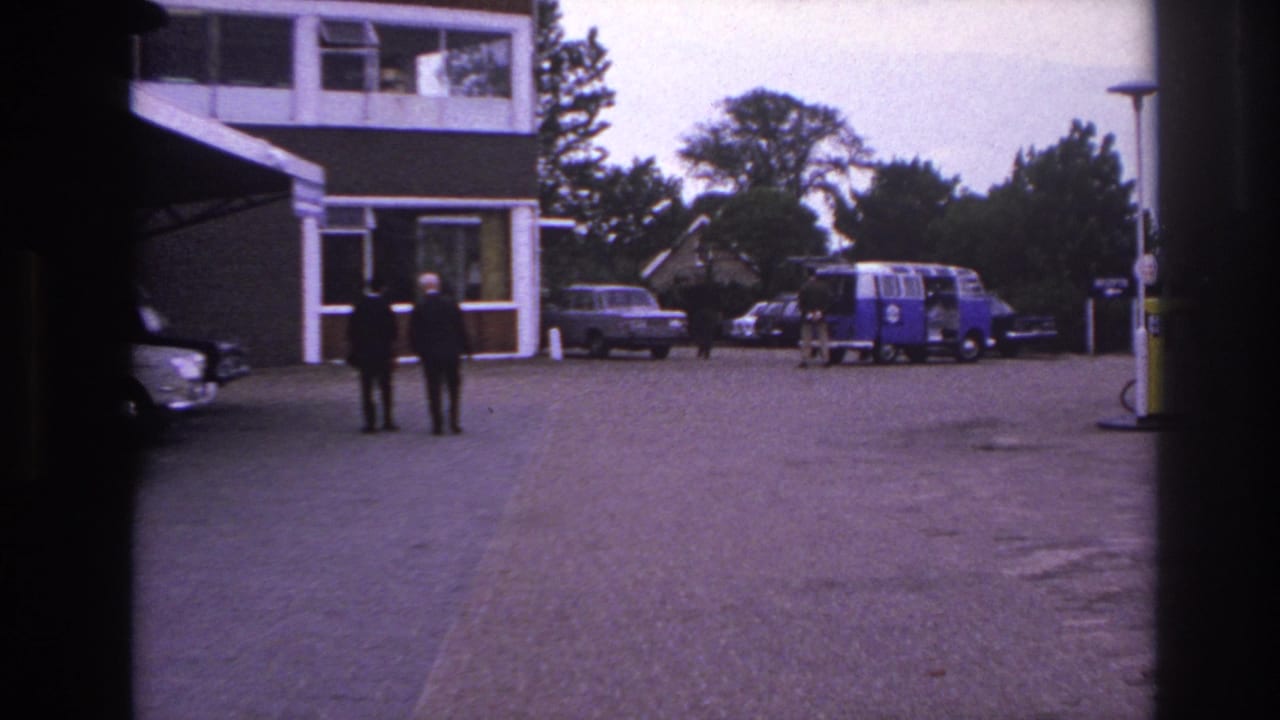 Two men in dark suits cross a Paris parking area in 1967, with period cars, trees, and a blue-and-white van in the background.