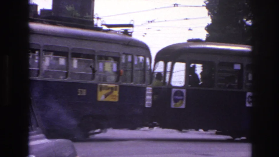 1967 Paris street frame with two dark buses passing close to camera, overhead wires, and trees behind them.
