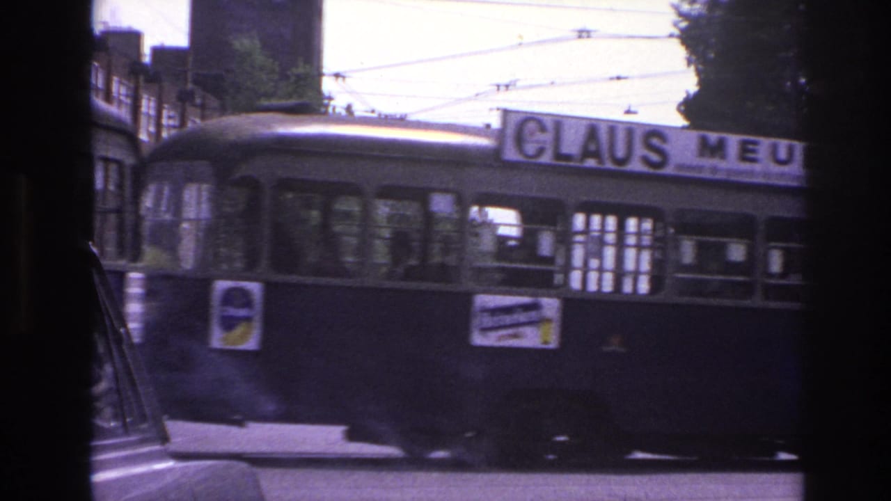 1967 Paris street view showing a trolley or bus moving past overhead cables, buildings, and a tree-lined intersection.