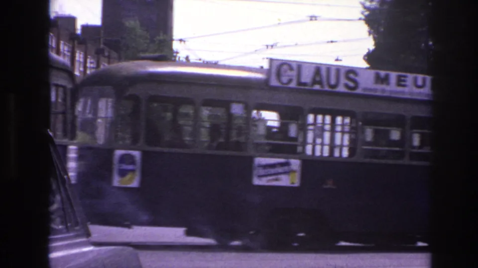 1967 Paris street view showing a trolley or bus moving past overhead cables, buildings, and a tree-lined intersection.