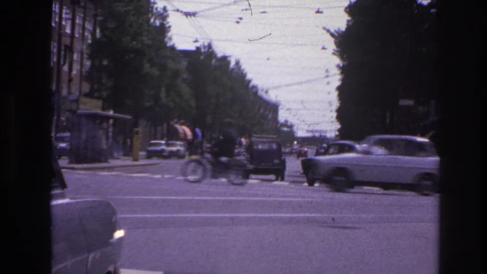 1967 Paris intersection with a cyclist, period cars, crosswalk stripes, overhead wires, and tree-lined streets seen from inside a vehicle.