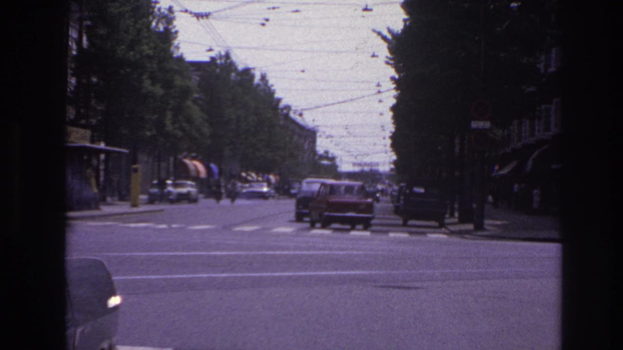 Classic cars travel along a broad Paris boulevard in 1967, with overhead wires, roadside trees, and dark window framing visible.