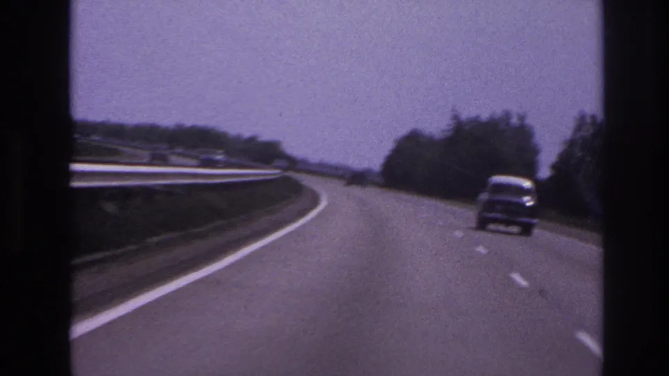 A classic car moves along a curving road in archival 8mm footage, with a guardrail, trees, and window-frame edges visible.