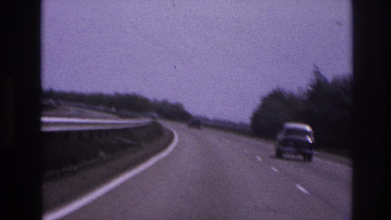 1967 Paris-area highway view from a moving vehicle, showing a classic car ahead on a curving road with guard rail and trees.