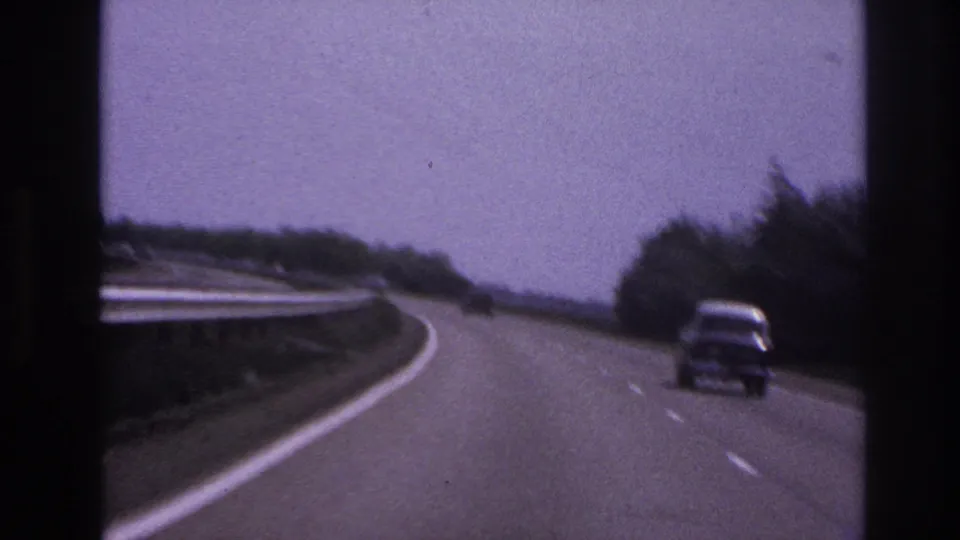 1967 Paris-area highway view from a moving vehicle, showing a classic car ahead on a curving road with guard rail and trees.