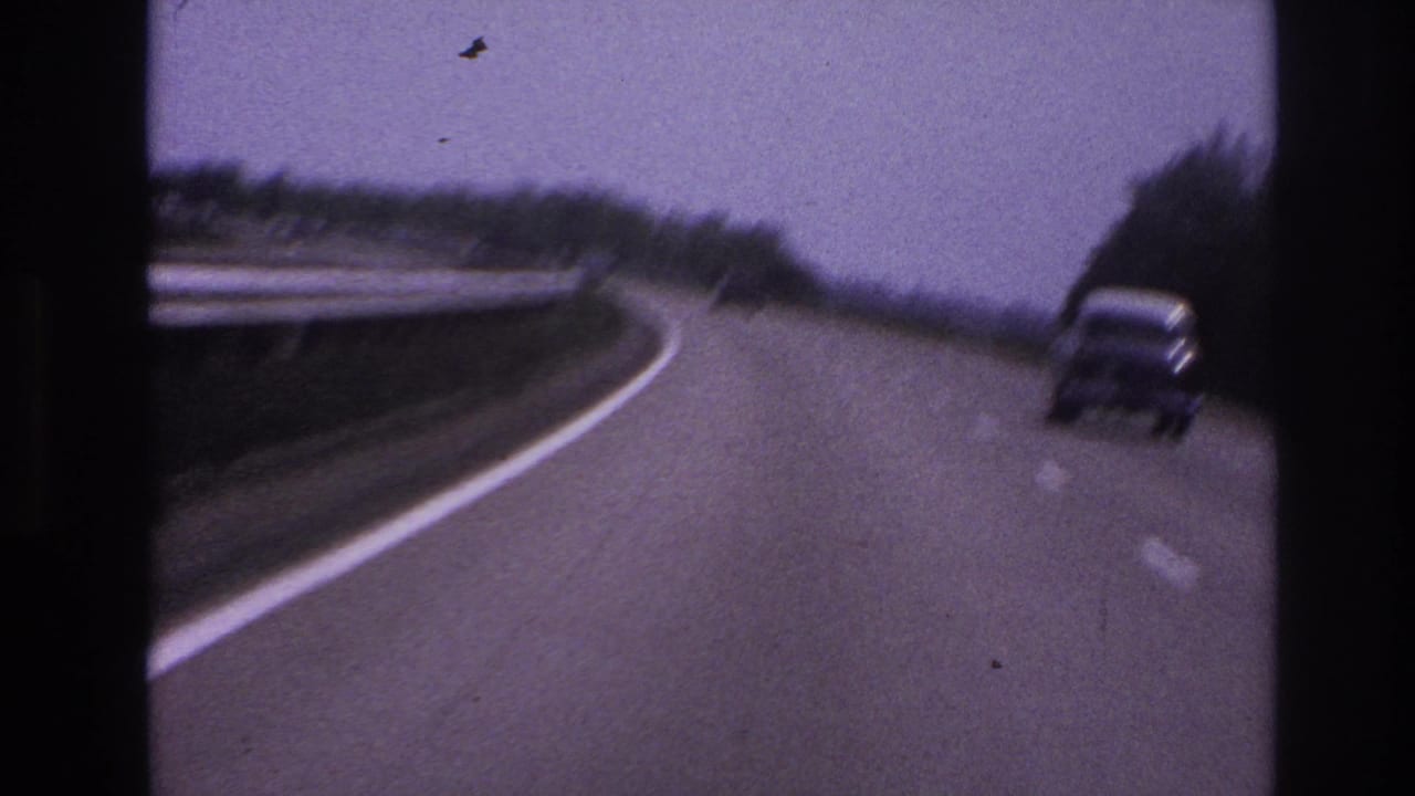 Highway bend with a car near the right lane, roadside guard rail, trees, and visible film grain.